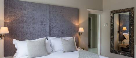 A neatly made bed with white linens and multiple pillows, a bedside table with a lamp, and a large mirror on the wall.