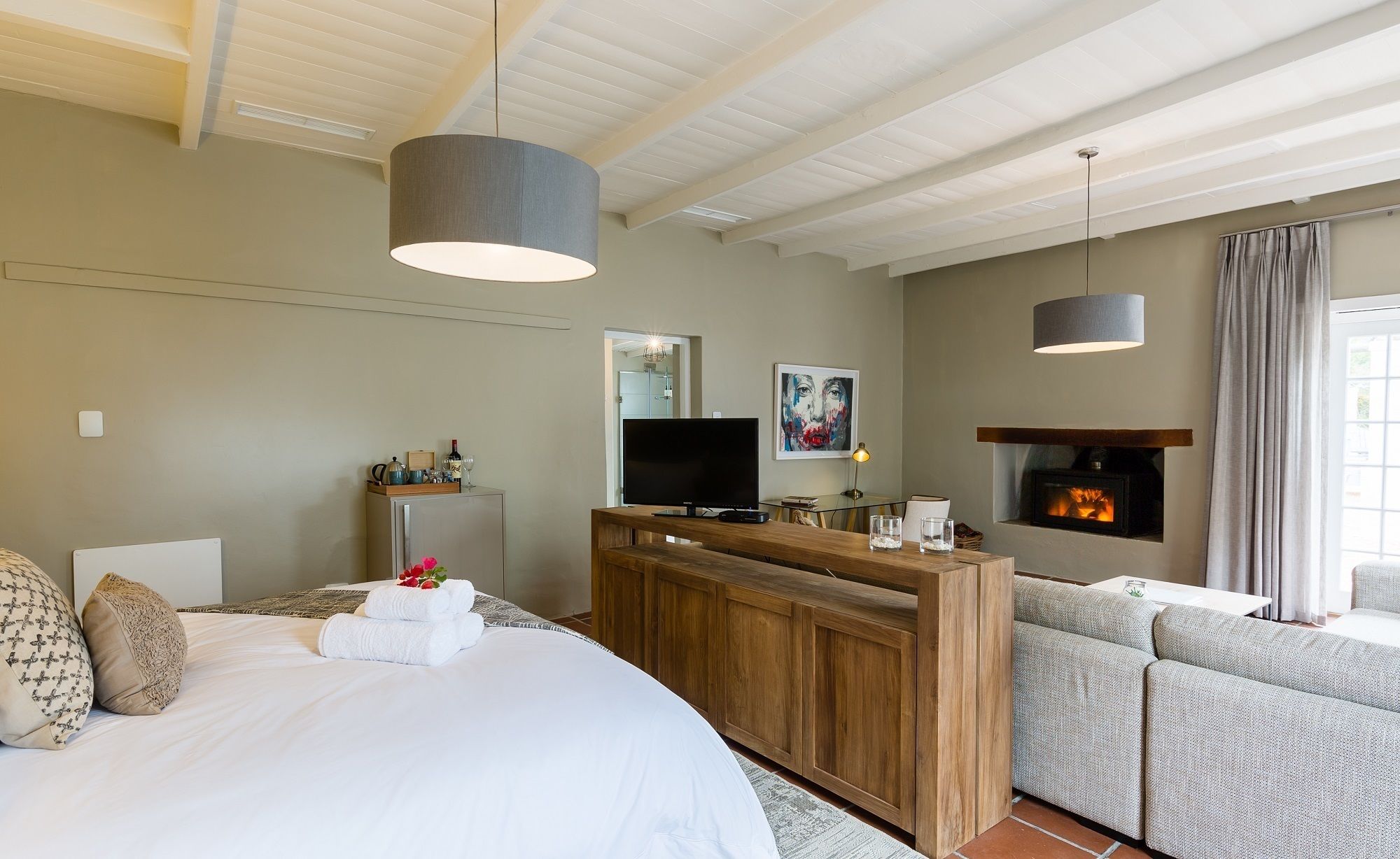 A modern hotel room with a large bed, a wooden console table, a TV, a fireplace, and a seating area with a sofa and armchairs.