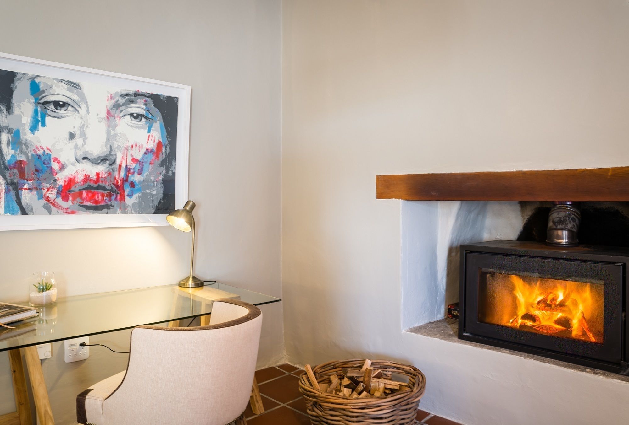 Een kamer met een glazen bureau, een stoel, een ingelijste kunstwerk, een open haard met gloeiende blokken hout, en een mand vol hout.