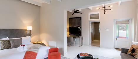 Habitación de hotel moderna con una cama grande, un escritorio, una silla y un sofá. Cuenta con un ventilador de techo, un televisor montado en la pared y una puerta que da a un baño.