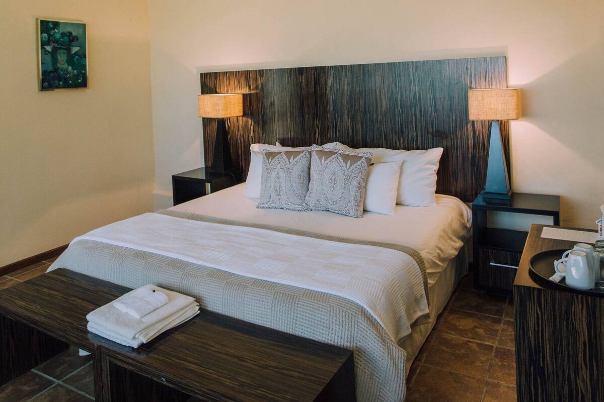A neatly arranged hotel room with a large bed, two bedside lamps, a wooden bench with towels, and a small table with a tray of mugs.