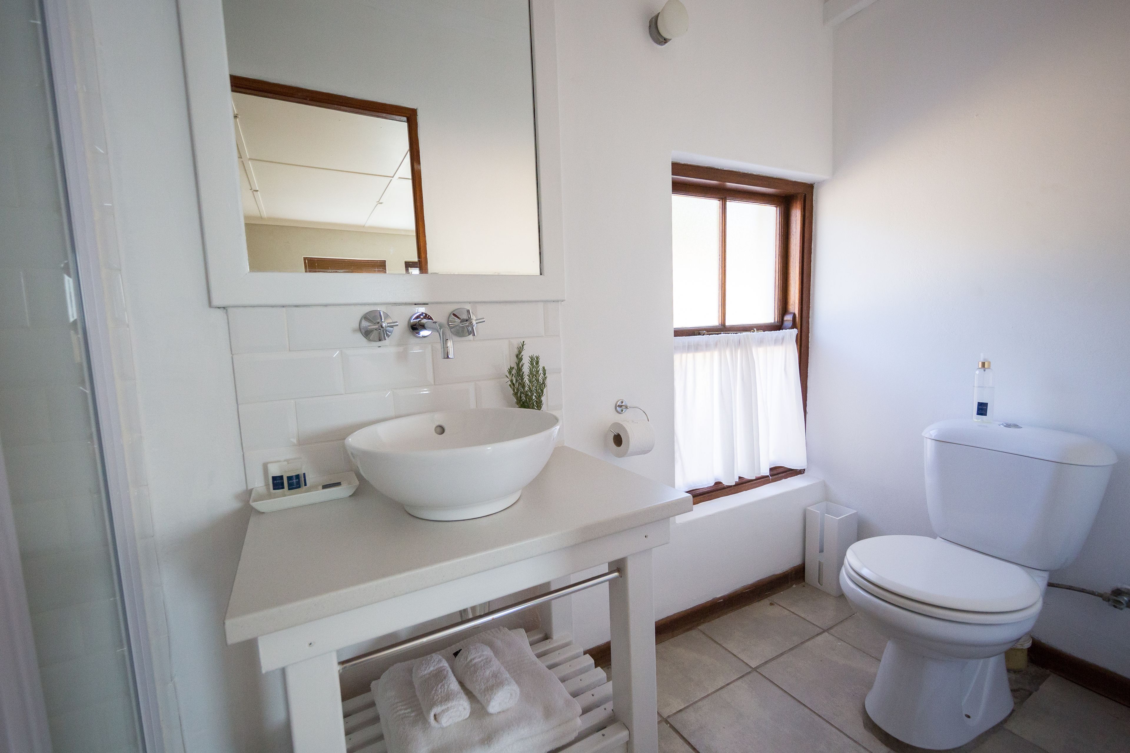 Un baño con lavamanos blanco, espejo e inodoro. Hay una ventana con cortinas y un perchero con toallas.