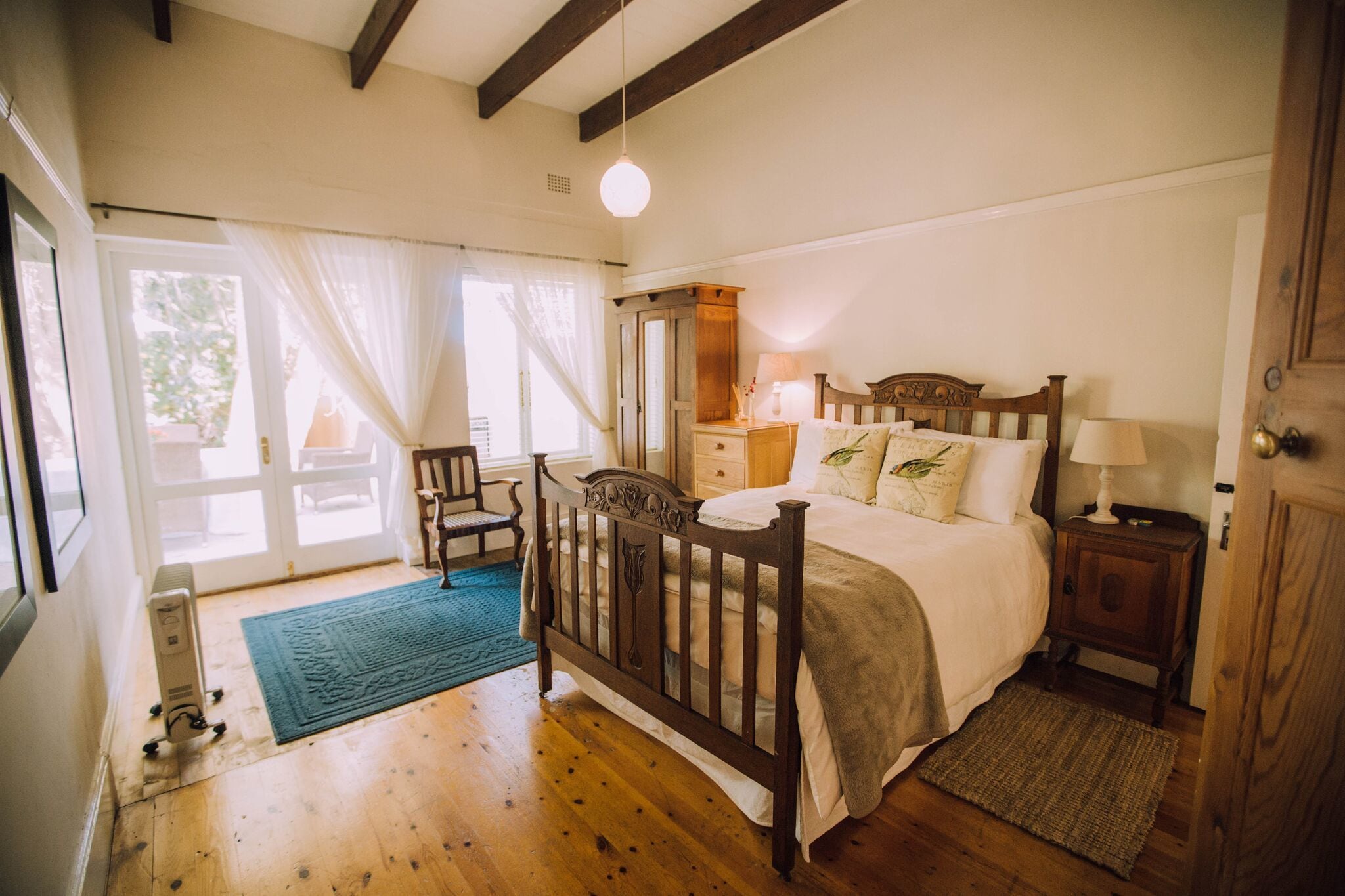 Un dormitorio con una cama de madera, una mesita de noche, una silla y una ventana con cortinas traslúcidas.