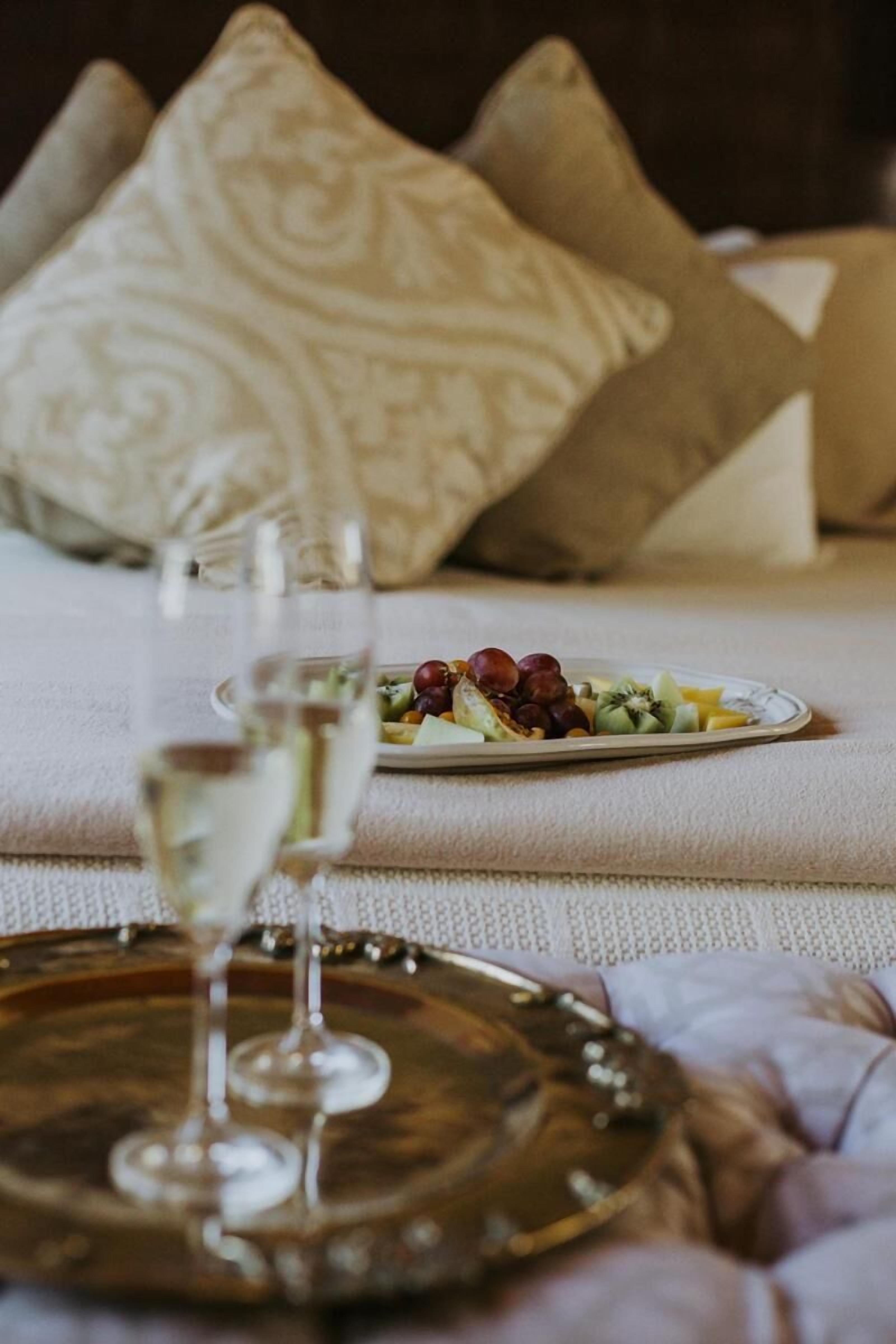 Una mesa de comedor con un plato de fruta y una copa de vino blanco, frente a una cama con almohadones estampados.