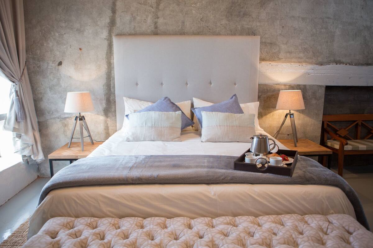 A bedroom with a large bed, two bedside tables with lamps, a tray with a teapot and cups, and a view of a fireplace.