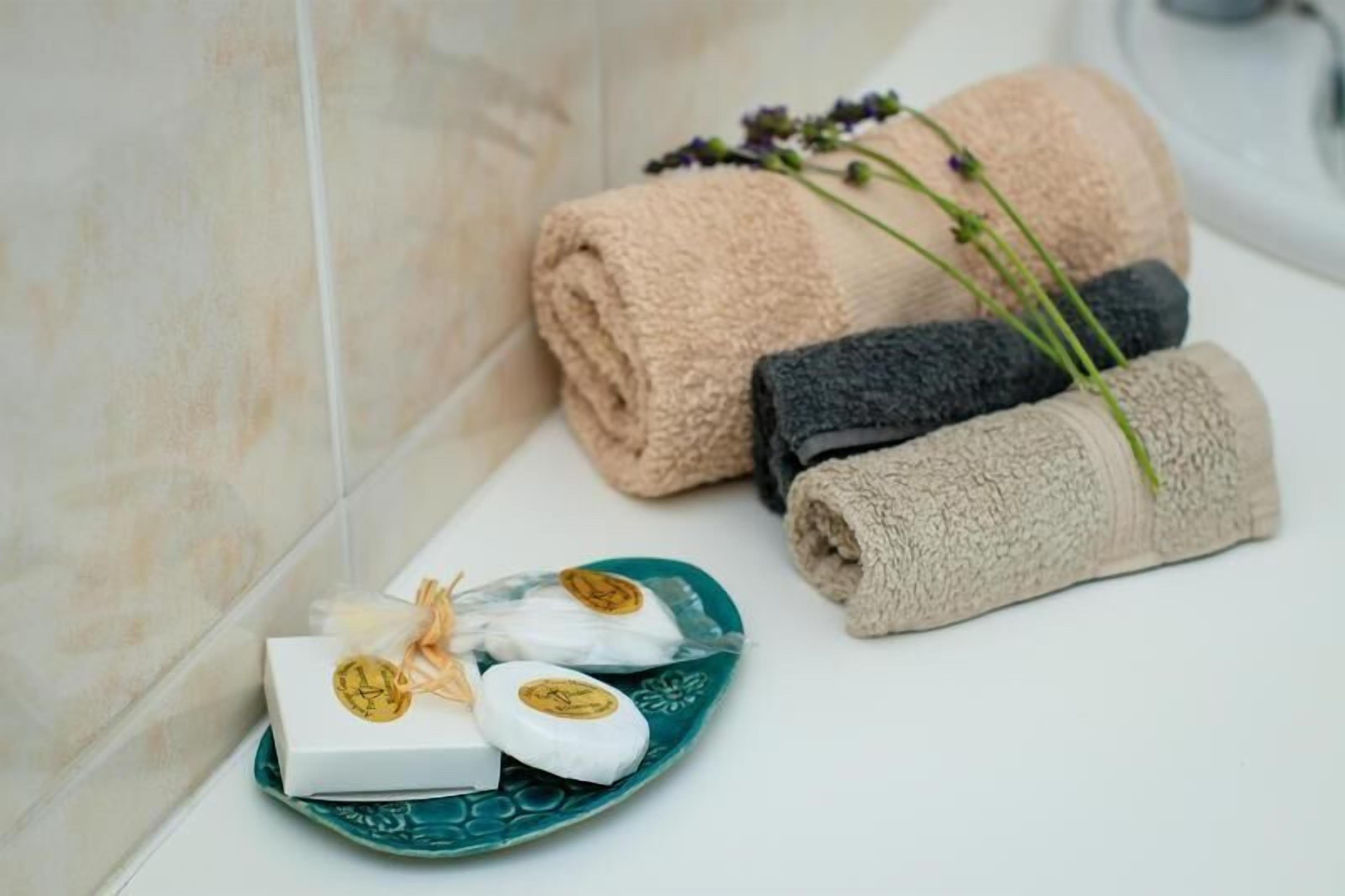 A bathroom with rolled towels in beige, black, and gray, a green plate with soap, and a sprig of lavender.