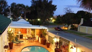 A swimming pool with a curved edge, surrounded by a patio area with chairs and tables, and a covered outdoor seating area. - Anchorage Guest House (Gqeberha (Port Elizabeth))
