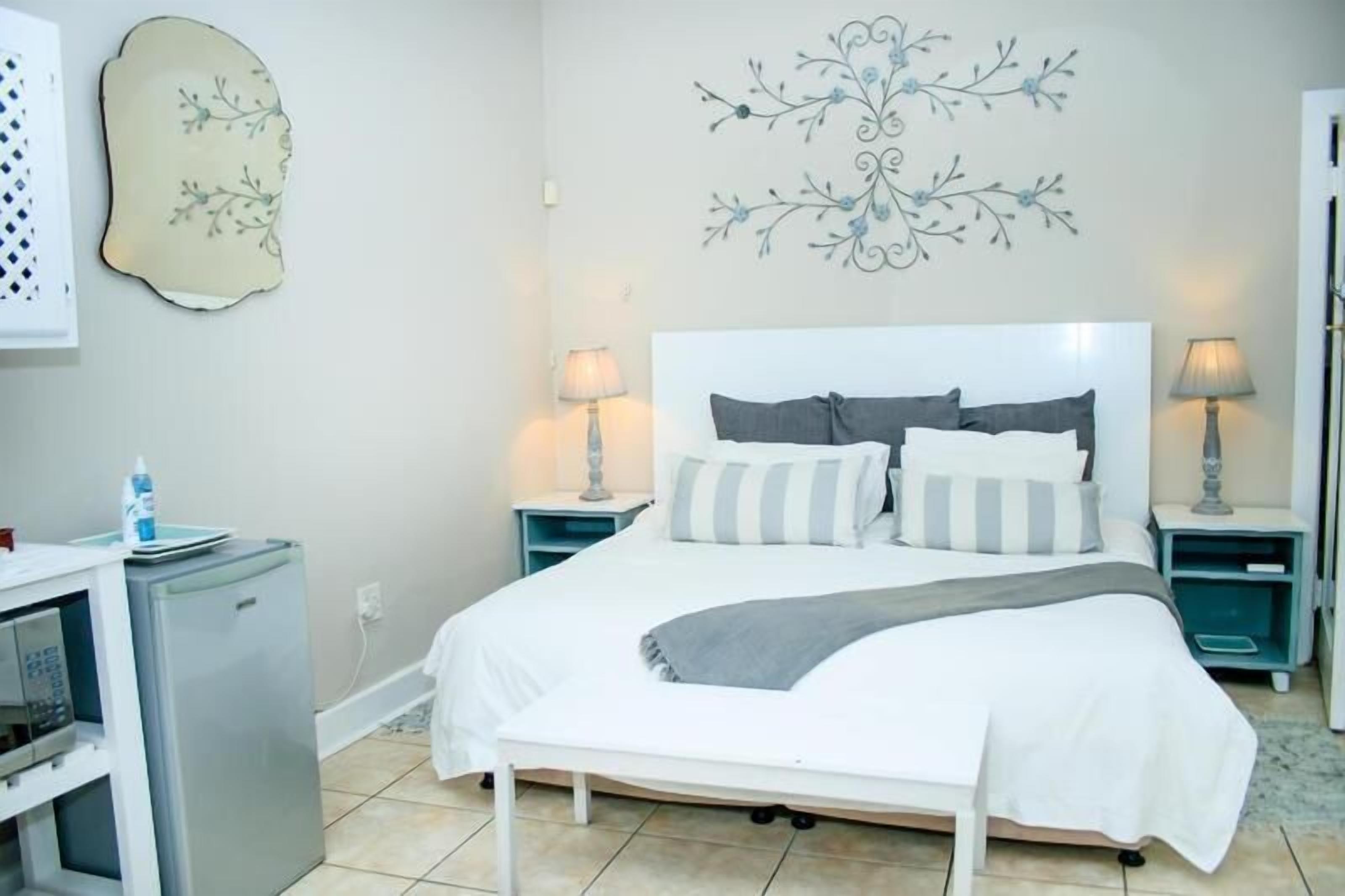 A neatly arranged bedroom with a bed, two bedside tables with lamps, a small refrigerator, and decorative wall art.