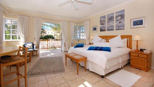 A hotel room with a bed, a desk, a chair, a small table, a nightstand, and a ceiling fan. - Anchorage Guest House (Gqeberha (Port Elizabeth))