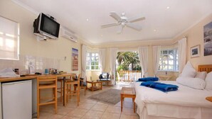 A hotel room with a bed, a desk, a television, a ceiling fan, and a view of the outdoors. - Anchorage Guest House (Gqeberha (Port Elizabeth))