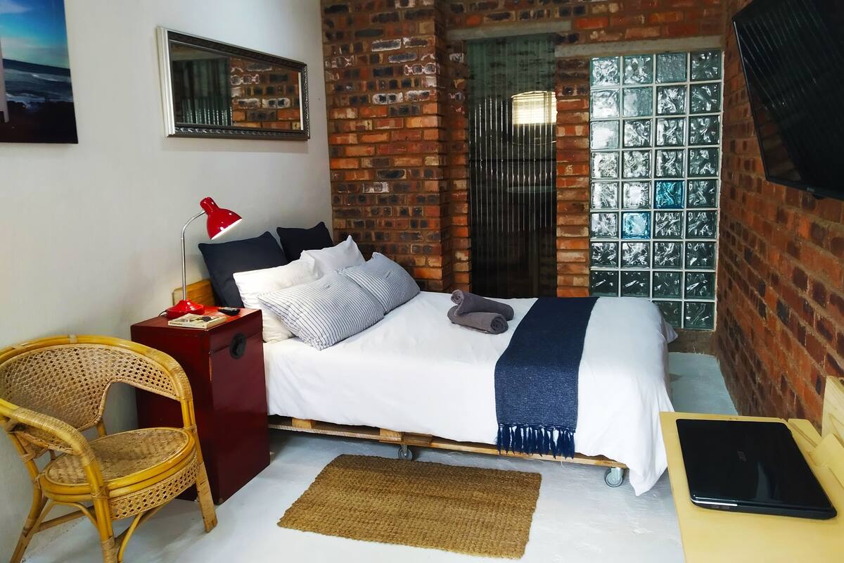 A bedroom with a brick wall, a bed with white bedding, a red lamp, a wicker chair, and a tablet on a wooden table.