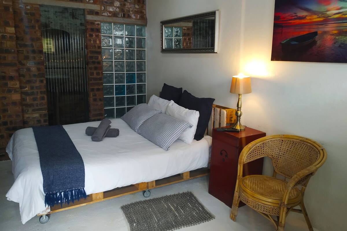 A neatly made bed with pillows and a blanket, a bedside table with a lamp, a wicker chair, and a painting on the wall.