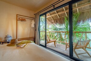 A hotel room with a large bed, a wooden nightstand, a hat, and a book. There is a balcony with wicker chairs and a thatched roof.