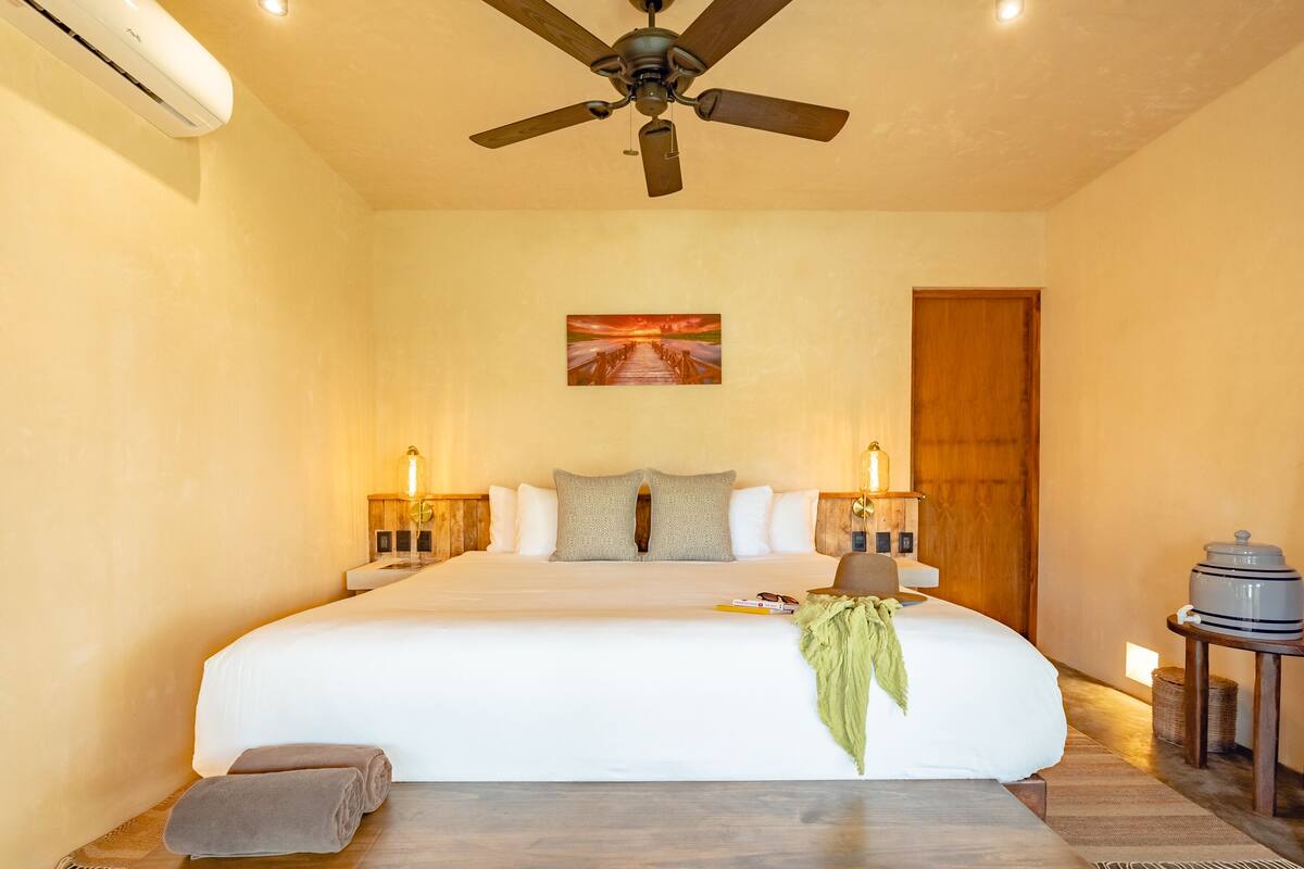 A large bed with white linens, a ceiling fan, and a painting of a sunset on the wall.