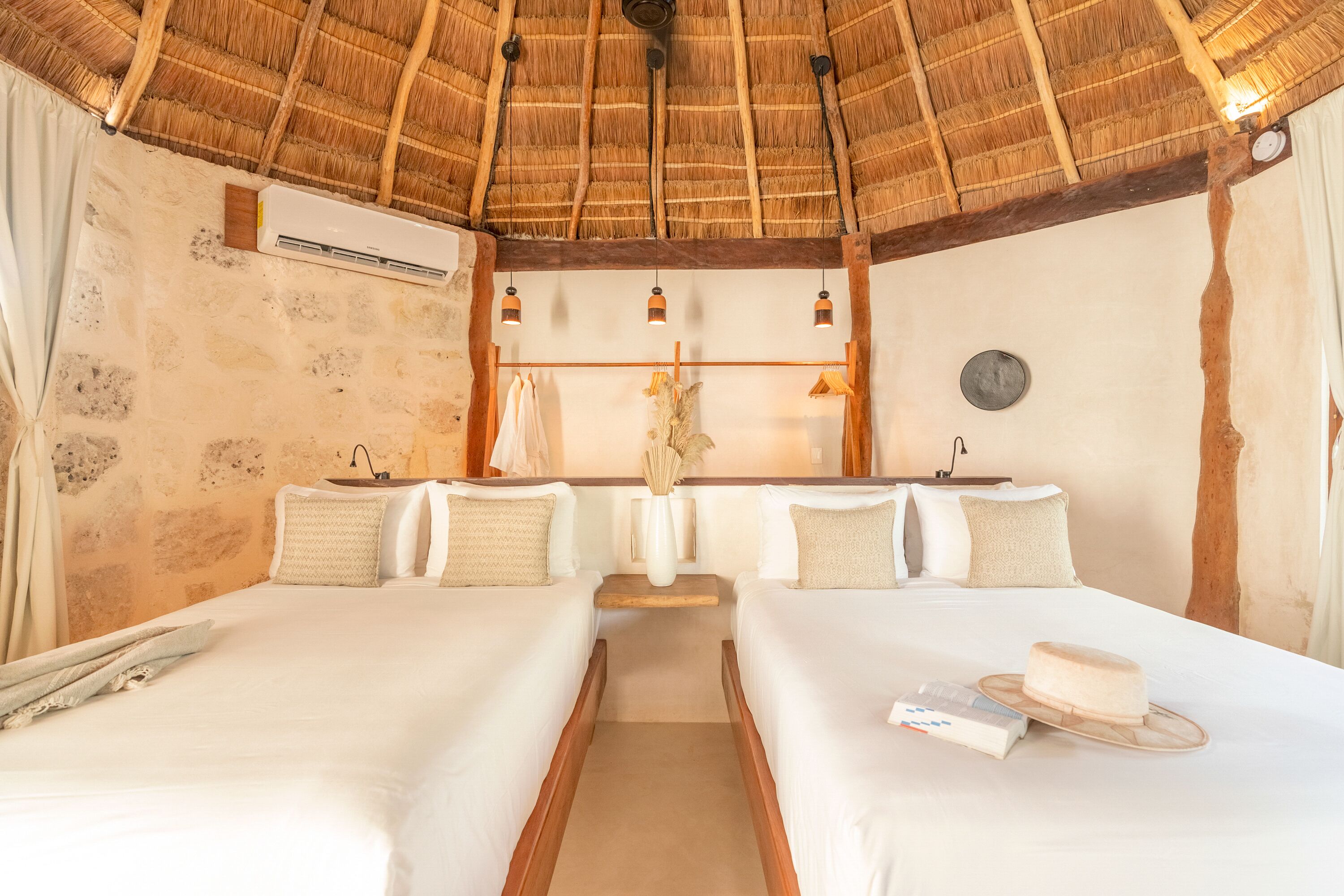 A room with two beds, a thatched roof, a white air conditioner, and a small shelf with a vase and a clock.