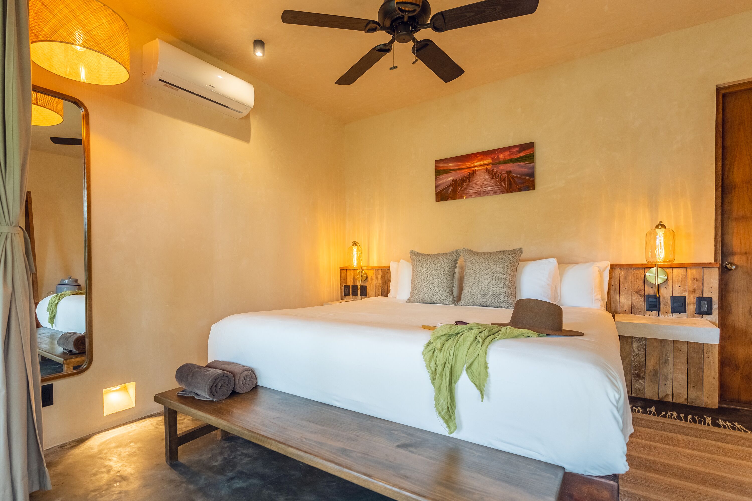 A hotel room with a large bed, a ceiling fan, an air conditioning unit, a bedside table with a lamp, and a painting on the wall.