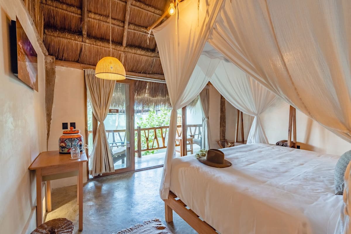 A room with a bed, a table, a hanging light, and a view of the outdoors.