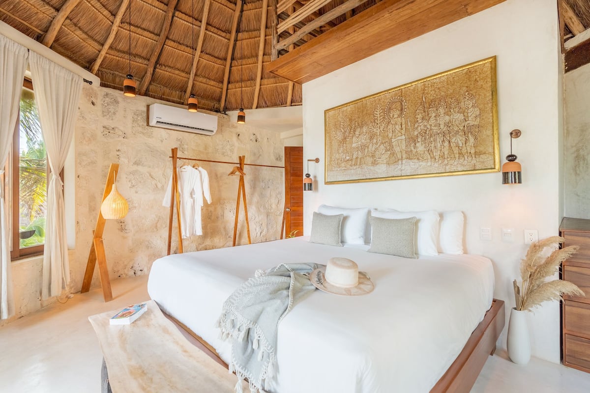 A bedroom with a large bed, a wooden ceiling, an air conditioning unit, a wall-mounted painting, and a bedside lamp.