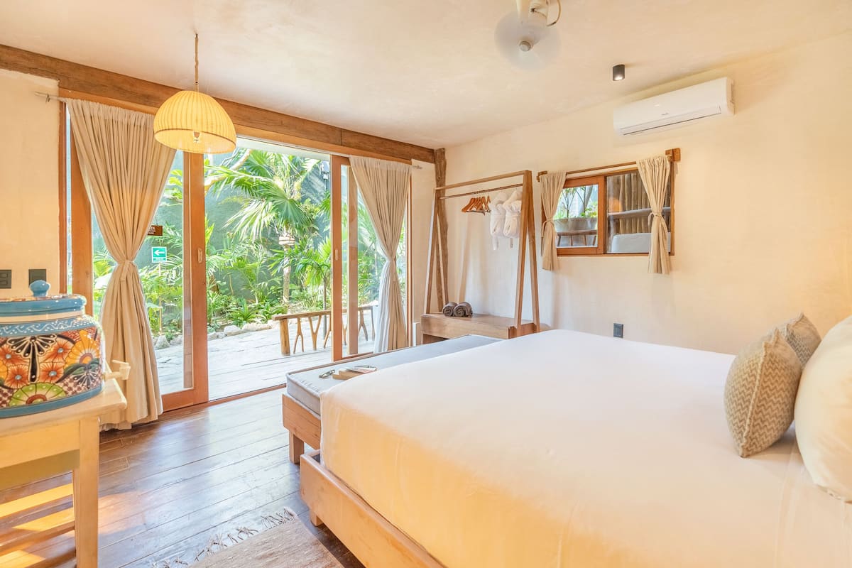 A bedroom with a large bed, wooden flooring, a small table with a colorful container, and a view of greenery through the window.