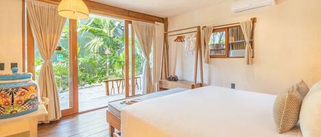 A bedroom with a large bed, wooden flooring, a small table with a colorful container, and a view of greenery through the window.