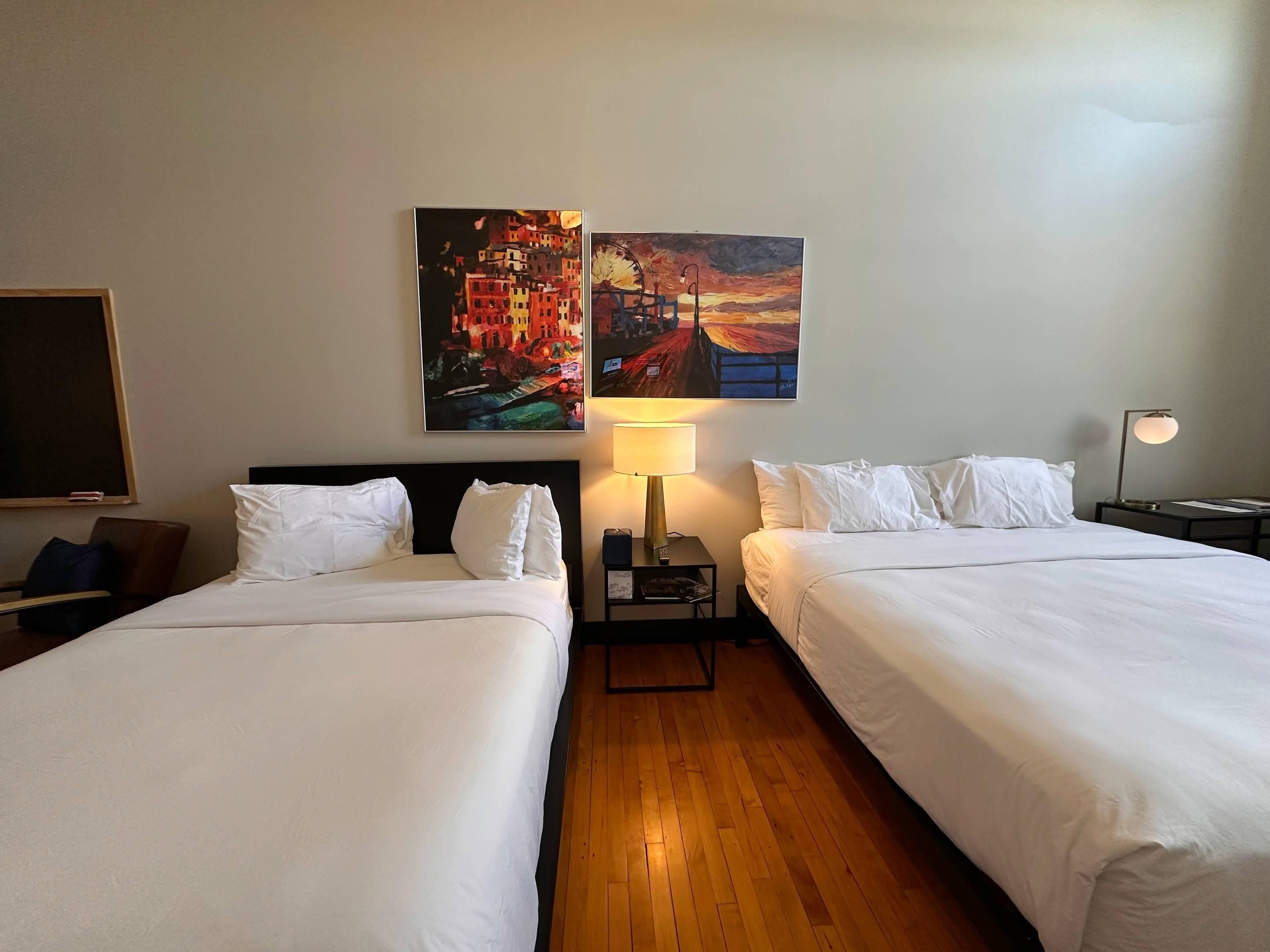 A hotel room with two beds, a nightstand with a lamp, and two paintings on the wall.