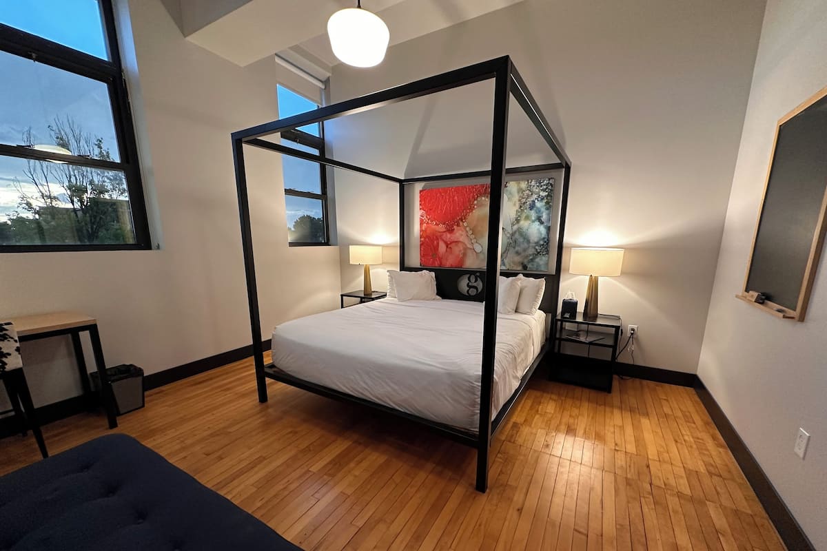 A four-poster bed with a modern design, two bedside tables with lamps, a small desk with a chair, and a large window with a view of trees.