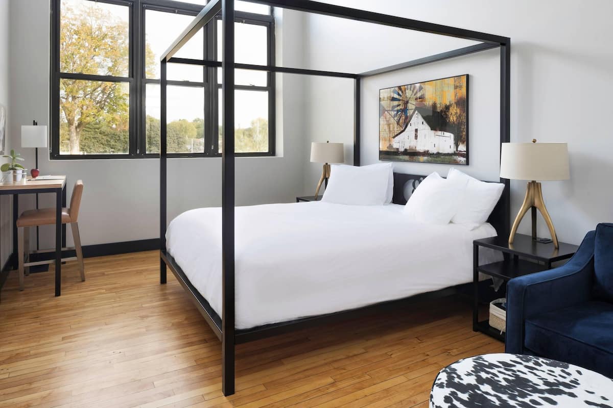 A modern bedroom with a four-poster bed, a desk, a chair, a lamp, and a painting on the wall.