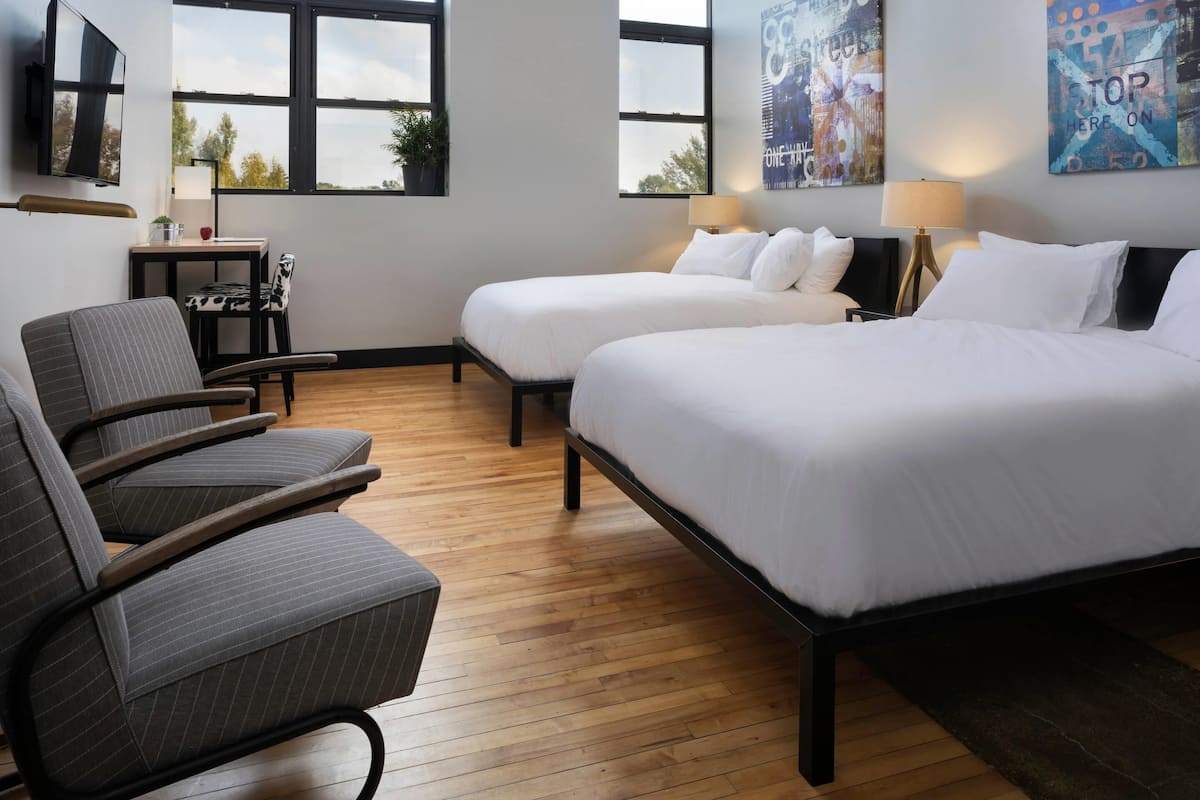 A hotel room with two beds, a desk, a chair, and large windows.