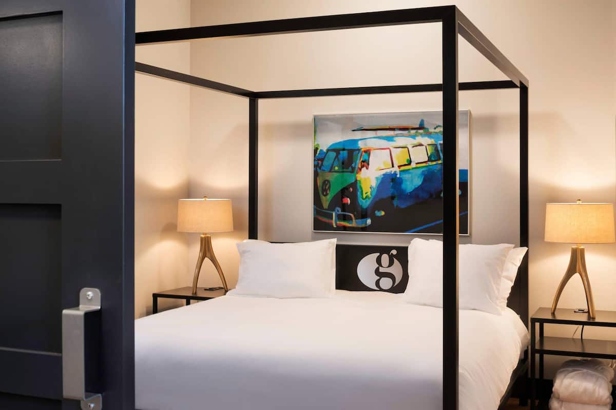 A four-poster bed with white bedding, two bedside tables with lamps, a painting on the wall, and a bench at the foot of the bed.