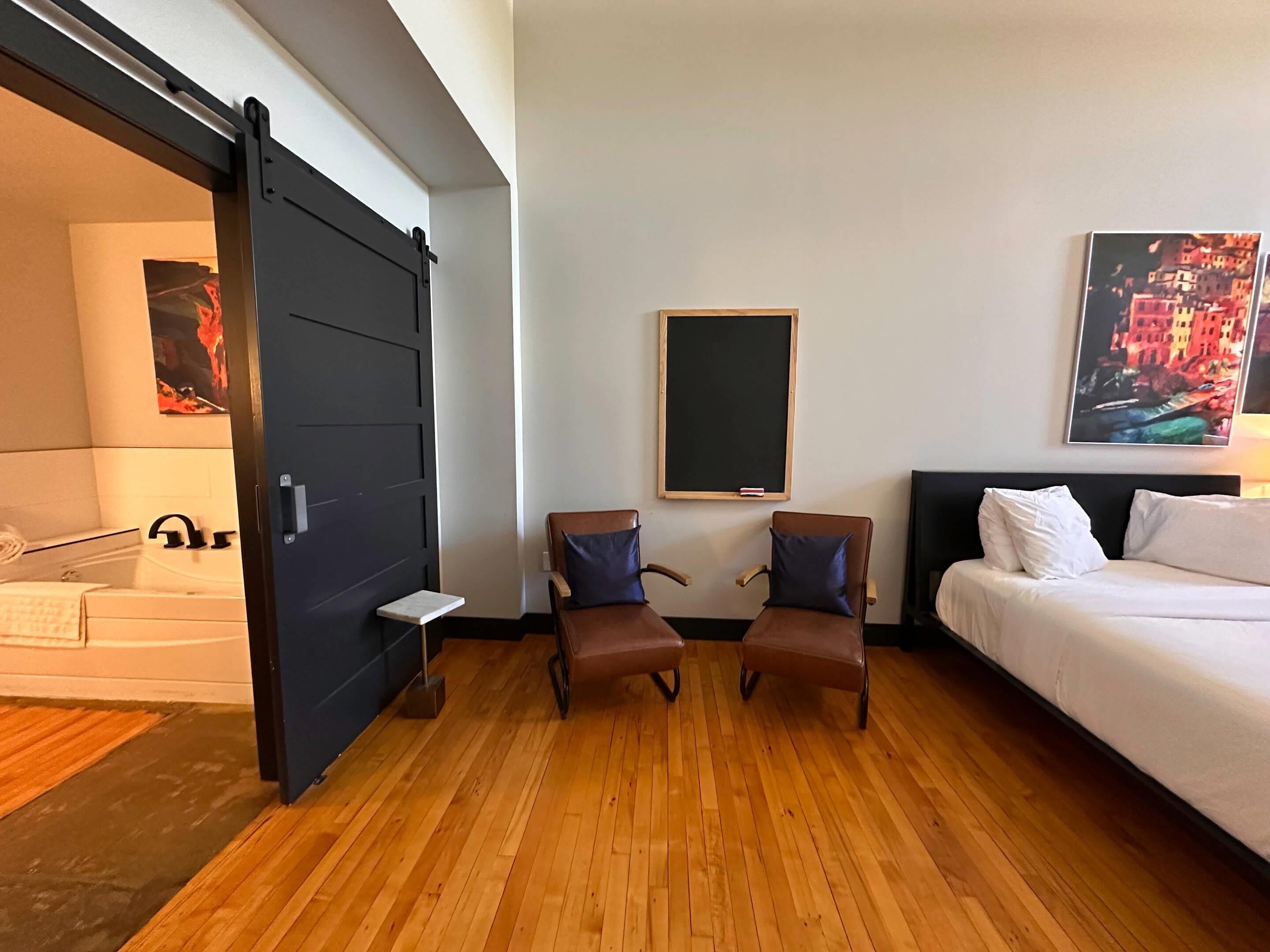 A hotel room with a bed, two armchairs, a small table, a blackboard, and a painting on the wall.