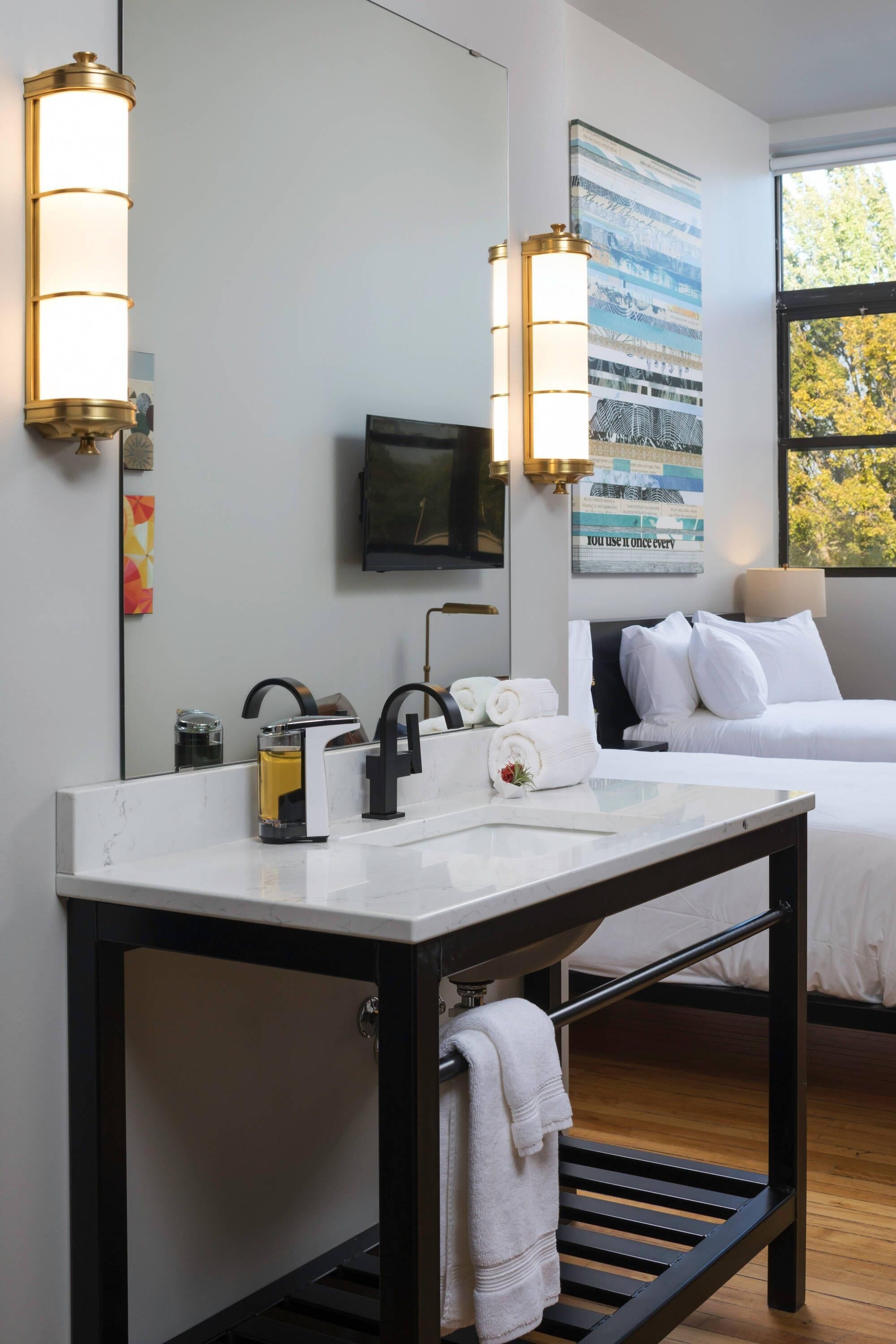 A hotel room with a bed, a bedside table with a lamp, a large mirror, and a wall-mounted TV.