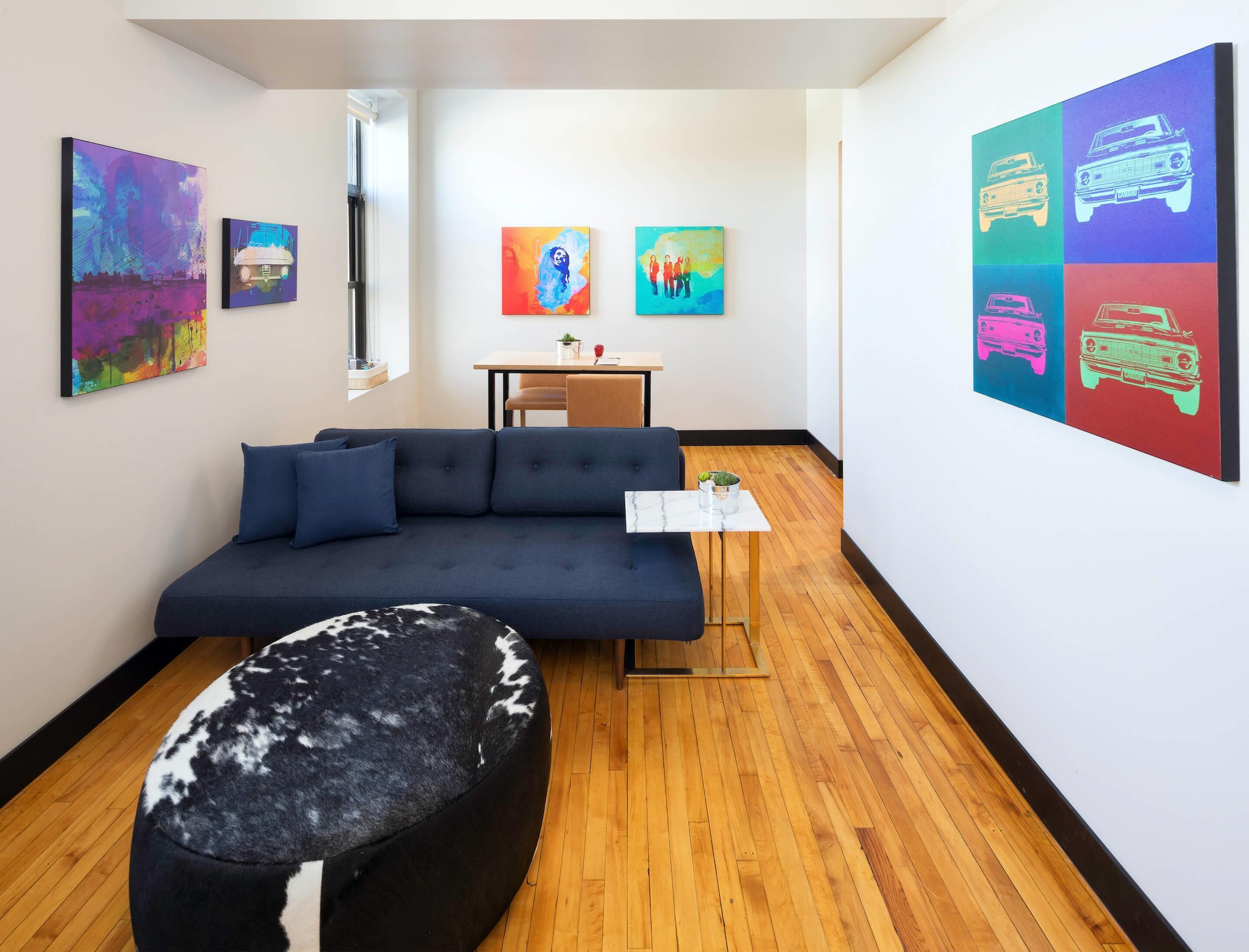 Un salon moderne avec un canapé bleu foncé, un pouf rond et une petite table sur laquelle est posée une plante. Les murs sont ornés de peintures abstraites aux couleurs vives, et une grande œuvre d’art pop représentant des voitures y est exposée.