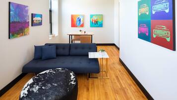 Un salon moderne avec un canapé bleu foncé, un pouf rond et une petite table sur laquelle est posée une plante. Les murs sont ornés de peintures abstraites aux couleurs vives, et une grande œuvre d’art pop représentant des voitures y est exposée.
