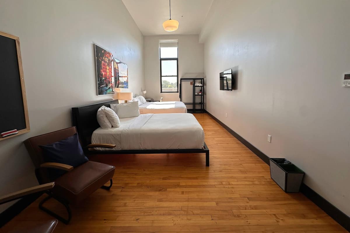 A hotel room with two beds, a desk, a chair, a TV, a painting, and a window.