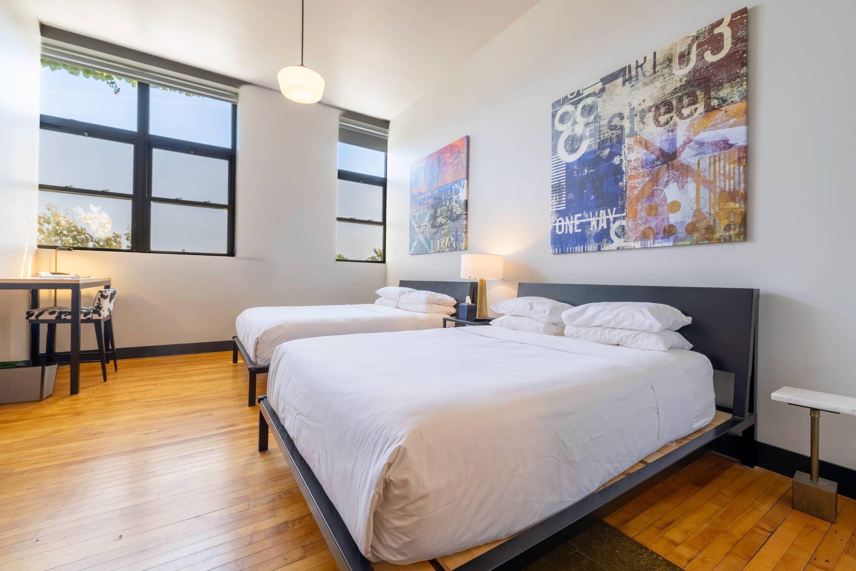 Une chambre d’hôtel avec deux lits, un bureau et une chaise. Elle dispose de grandes fenêtres et les murs sont décorés d’œuvres d’art.