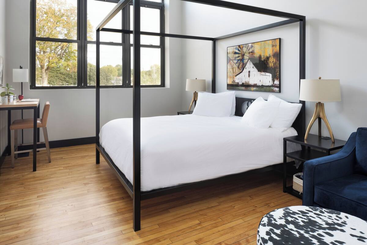A modern hotel room with a four-poster bed, a desk with a chair, a blue armchair, and a side table with a lamp.