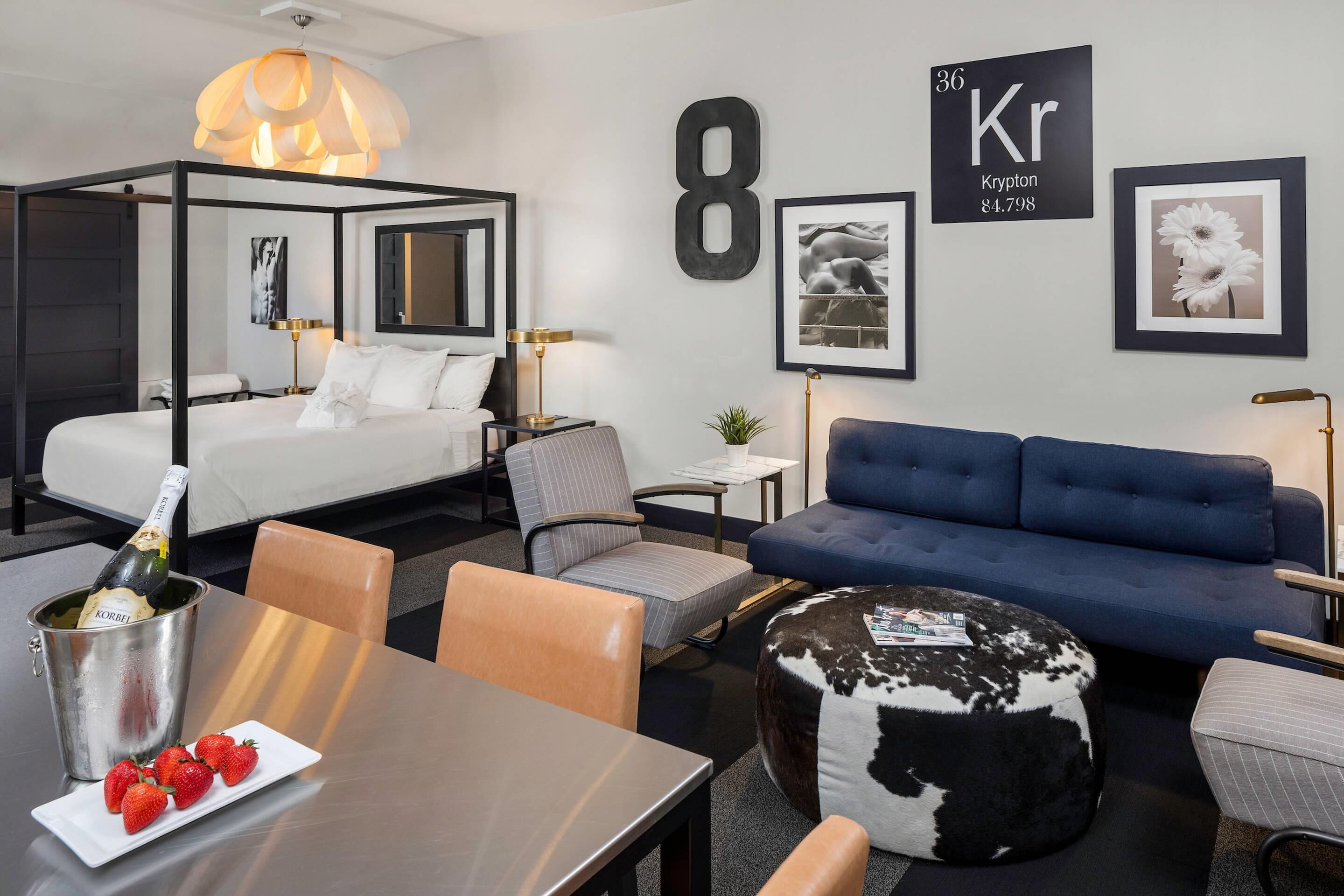 Une chambre d’hôtel moderne avec un lit à baldaquin, un canapé bleu, une table à manger décorée de fraises et une bouteille de champagne.