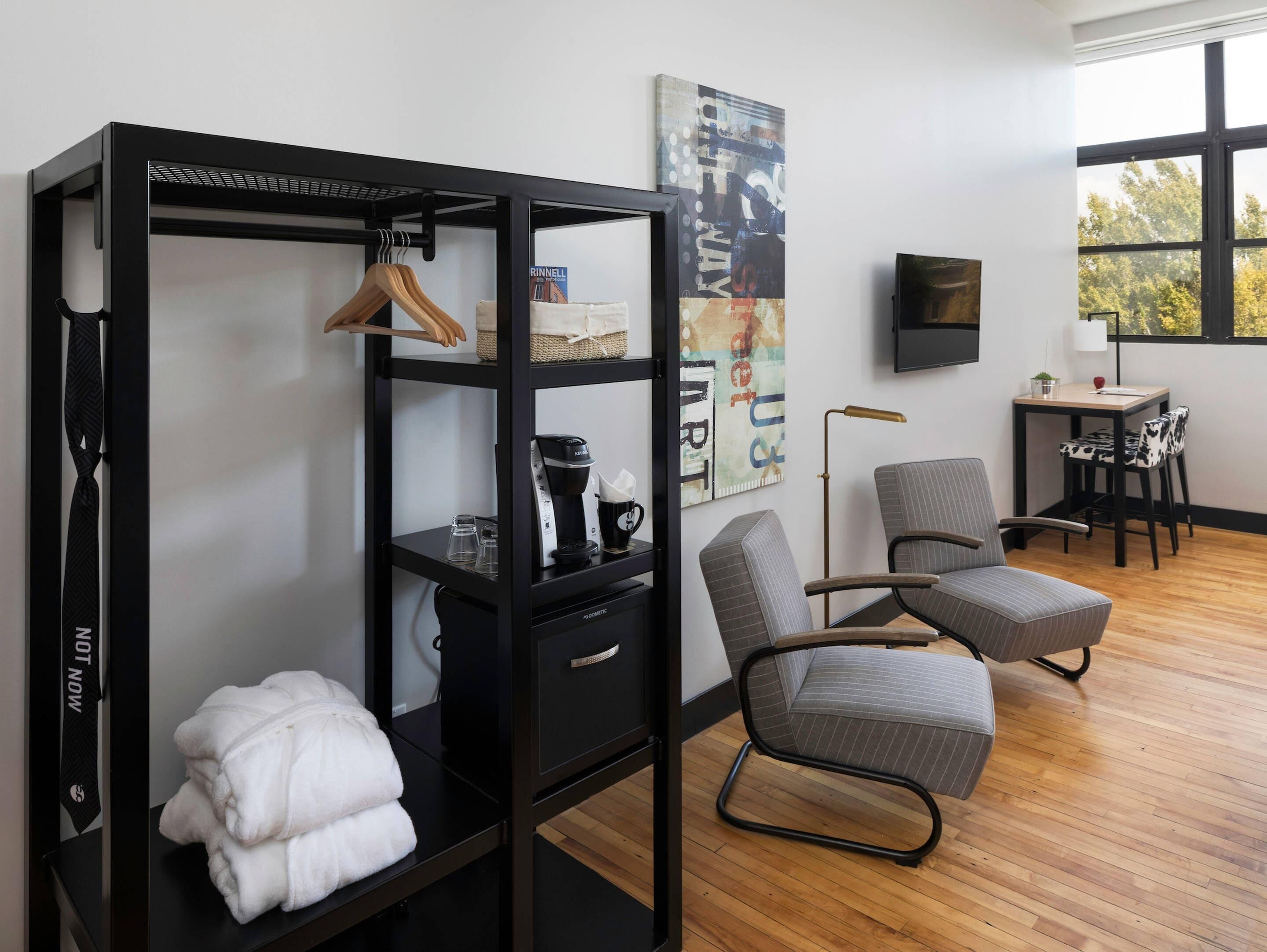 Ein modernes Zimmer mit einem schwarzen Metallregal, einem grau gepolsterten Sessel, einem Holztisch und einem großen Fenster mit Blick auf Bäume.