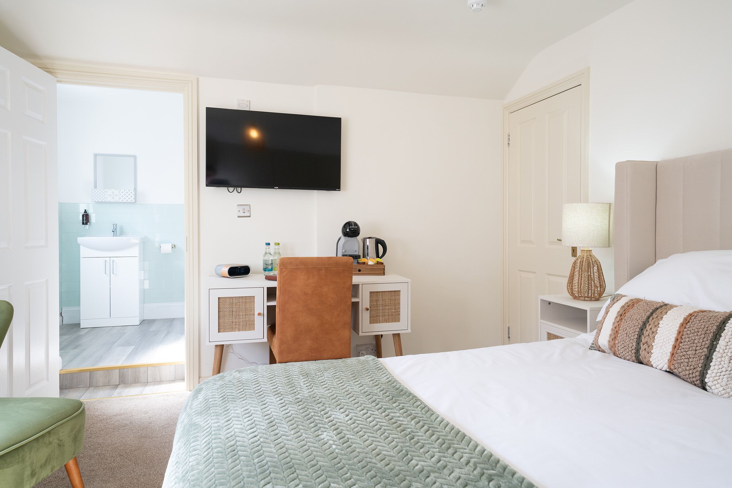 Quarto de hotel com cama, escrivaninha com cadeira, televisão, luminária e banheiro visível por uma porta aberta.