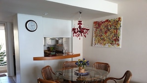 A dining area with a round glass table surrounded by wicker chairs, a kitchen area with white cabinets, a clock, and a colorful abstract painting on the wall. - Departamento Fragata (Algarrobo)
