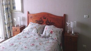 A bedroom with a wooden headboard, floral bedding, two bedside tables with lamps, and a mirror on the wall. - Departamento Fragata (Algarrobo)