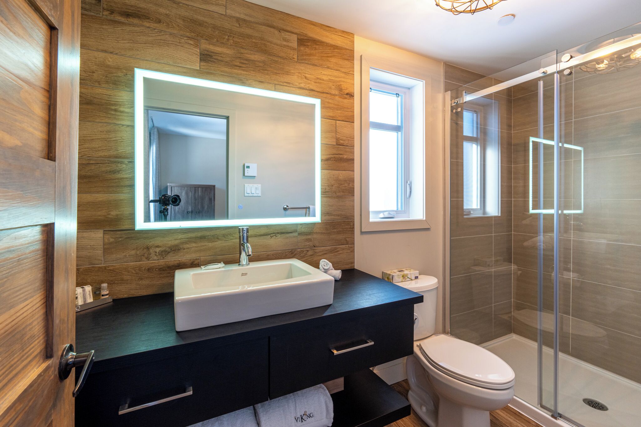 Un baño moderno con pared de madera, un espejo grande, un lavabo blanco, un mueble de tocador negro, un inodoro y una cabina de ducha de vidrio.