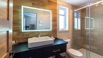 Un baño moderno con pared de madera, un espejo grande, un lavabo blanco, un mueble de tocador negro, un inodoro y una cabina de ducha de vidrio.
