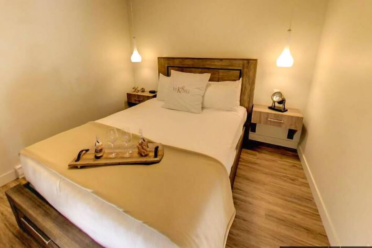 Un lit bien fait, avec une tête de lit en bois, une table de chevet sur laquelle se trouve une lampe, et un plateau avec des verres et une bouteille posé sur le lit.