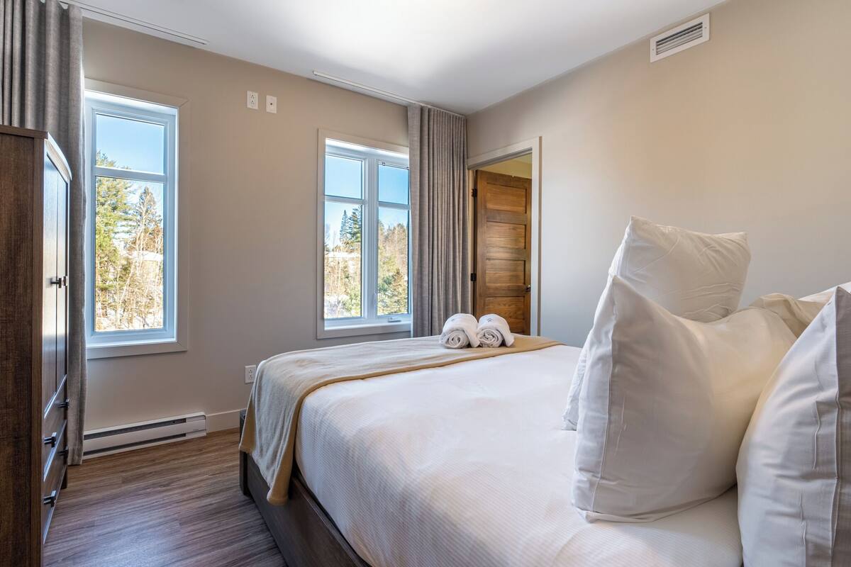 Une chambre à coucher avec un grand lit, deux fenêtres, une armoire en bois et une porte donnant sur une autre pièce.
