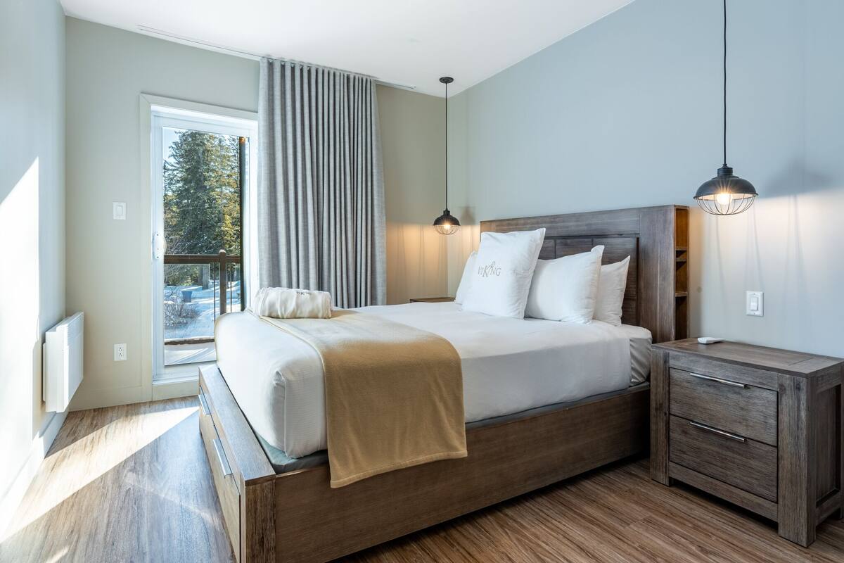 Une chambre à coucher avec un grand lit, une table de chevet, une commode, une fenêtre avec des rideaux et un luminaire suspendu.