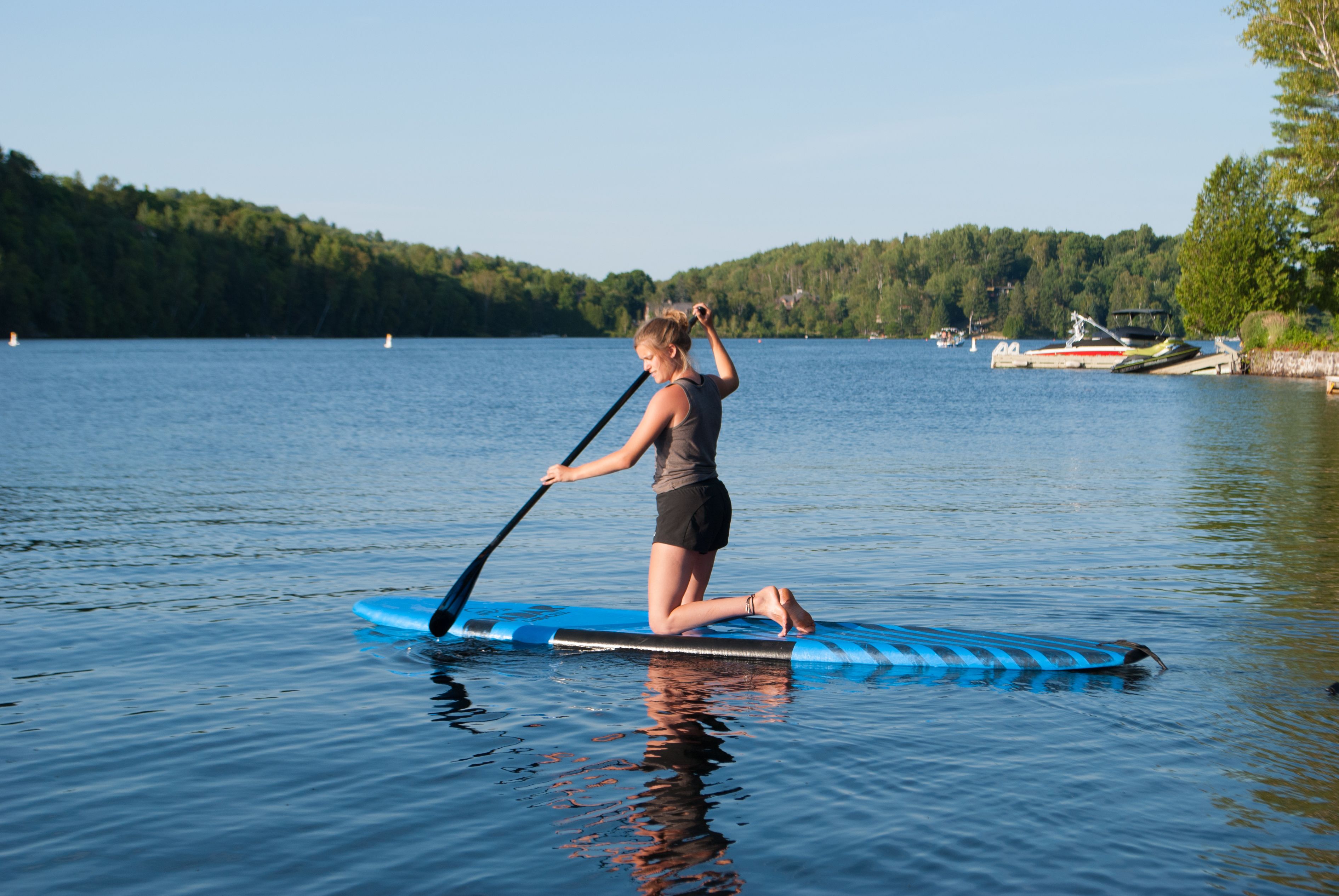Una persona haciendo paddleboard en un lago tranquilo, con barcos y árboles al fondo.