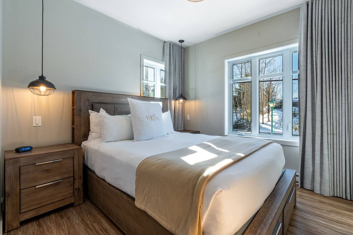 Un lit bien fait, avec une tête de lit en bois, une table de chevet avec une lampe, et une vue sur un paysage enneigé par de grandes fenêtres.
