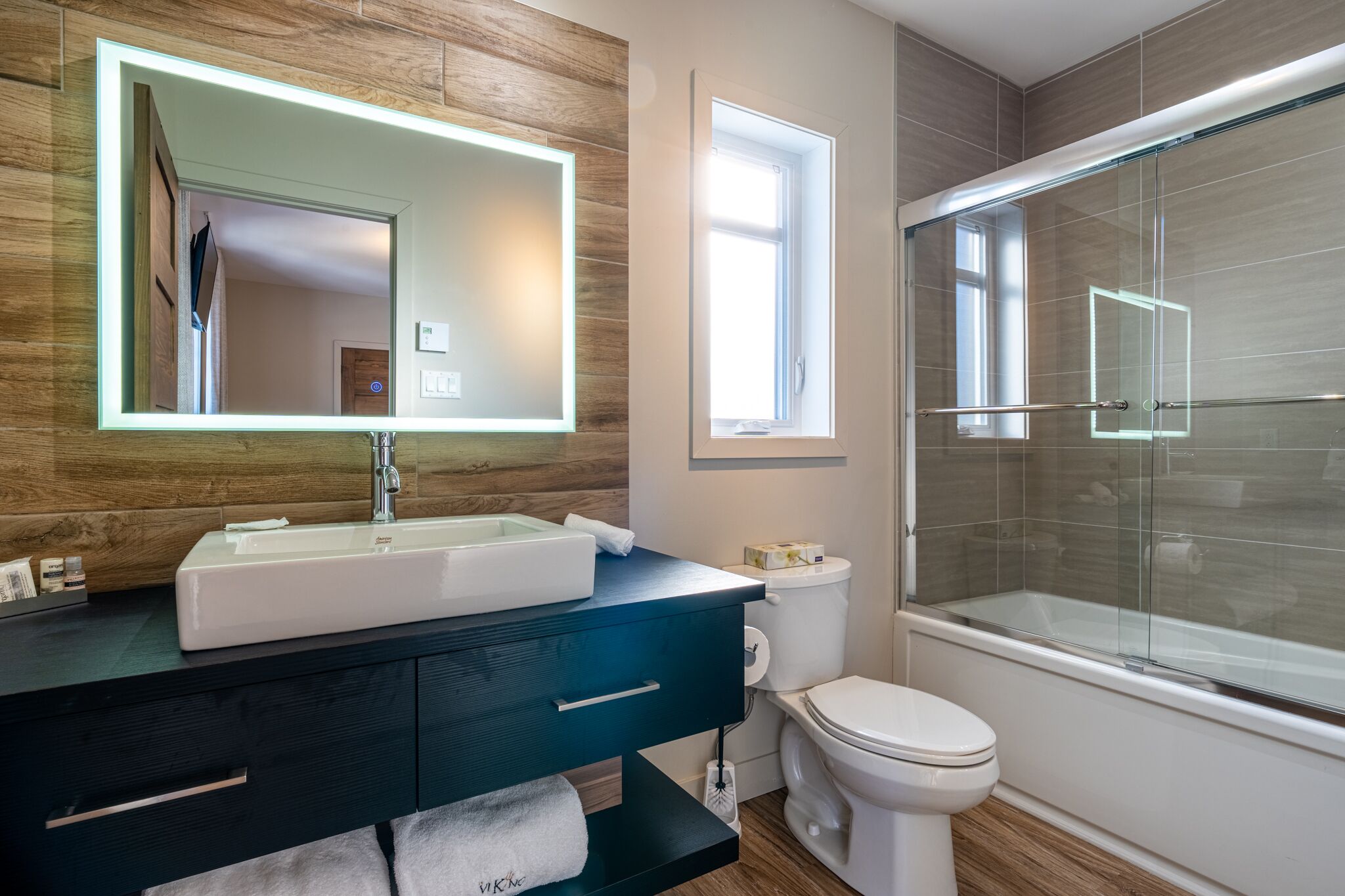 Un baño moderno con tocador de madera, un espejo grande, un lavabo blanco, inodoro y cabina de ducha con mampara de vidrio.