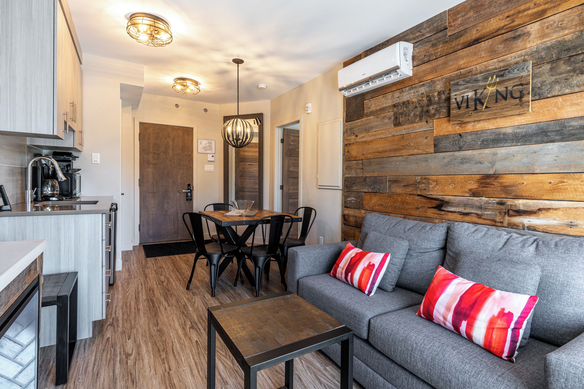 A modern kitchen with a built-in stove, a dining area with a wooden table and chairs, and a gray sofa with a red and white pillow.